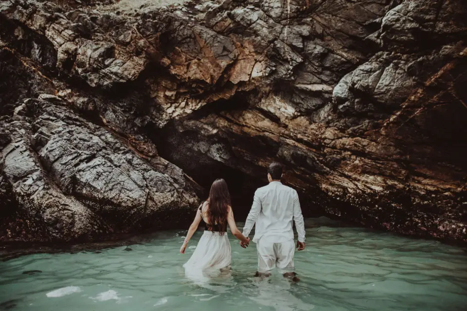 Rear view of a young couple holding hands while wading into clear sea water toward a rocky coastal cave formation.